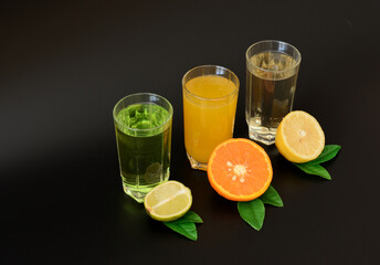 Three glasses with different citrus juices on a black background, next to ripe fruits.