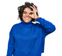 Young hispanic woman with curly hair wearing turtleneck sweater doing ok gesture with hand smiling, eye looking through fingers with happy face.