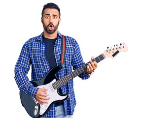 Young hispanic man playing electric guitar scared and amazed with open mouth for surprise,...