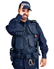 Young hispanic man wearing police uniform doing ok gesture with hand smiling, eye looking through fingers with happy face.