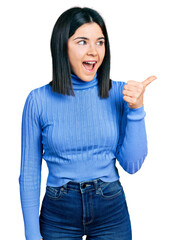 Young brunette woman with blue eyes wearing casual turtleneck sweater pointing thumb up to the side...