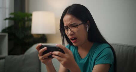 Portrait of Young Asian woman serious playing an online game frustrated with loss or failure sitting on sofa in living room at home.Gamer lifestyle concept.