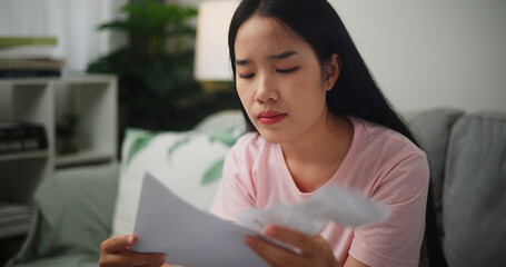 Portrait of Young Asian woman focused in calculating budget sitting on sofa at home holding paper taxes bills and reading financial information,Home finance