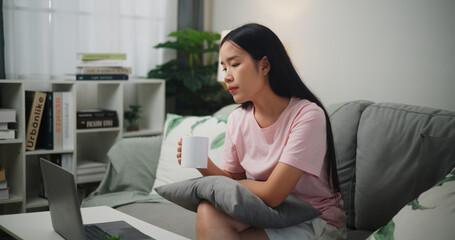 Portrait of Young asian woman thinking and using laptop while drink coffee on sofa at home,Home finance