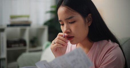 Portrait of Young Asian woman focused in calculating budget sitting on sofa at home holding paper...