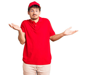 Young handsome man with curly hair wearing delivery uniform clueless and confused expression with...