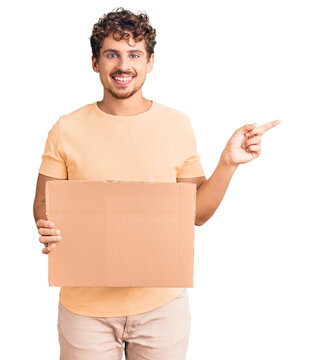 Young handsome man with curly hair holding blank cardboard empty banner smiling happy pointing with hand and finger to the side