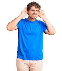 Young handsome man with curly hair wearing casual clothes trying to hear both hands on ear gesture, curious for gossip. hearing problem, deaf