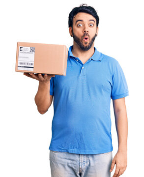 Young hispanic man holding delivery package scared and amazed with open mouth for surprise, disbelief face