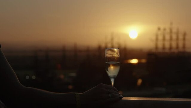 A woman steps onto the balcony to bid farewell to the sunset with a glass of champagne, tall masts of sailing ships in the distance at sea.