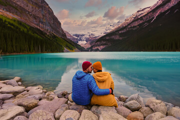 Lake Louise Banff National Park in the Canadian Rocky Mountains. A young couple of men and women sitting on a rock by the lake during a cold day in Autumn in Canada watching the sunset at the lake - obrazy, fototapety, plakaty