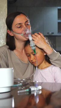 Mother and sad daughter at a table, woman in a mask inhaling medicine through a nebulizer. Mom gently hugs and soothes her daughter. Vertical video.