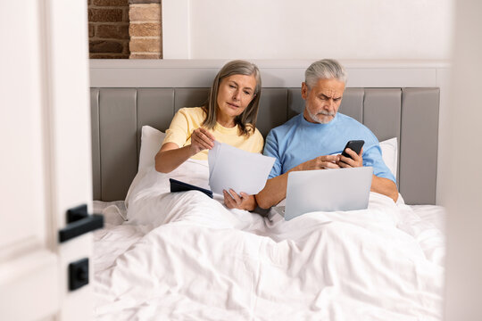 Mature Senior In Bed With Their Digital Gadgets Working From Home.