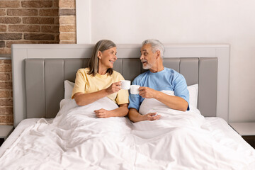Elderly couple sitting on bed enjoying coffee in cozy bedroom, radiating happiness and love. Man and woman relaxing in bed room drinking coffee in the morningin.