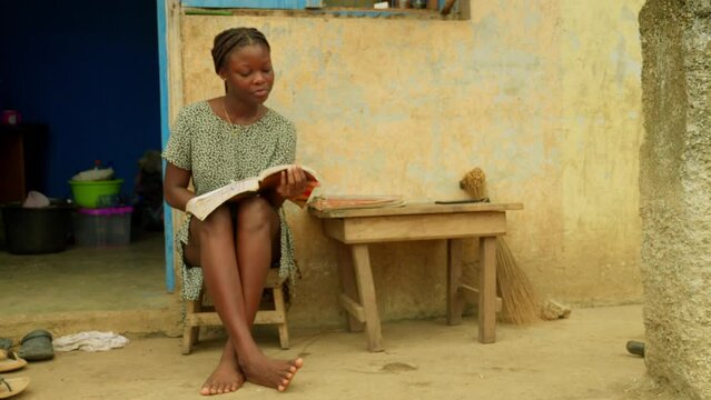 In front of a house, a young woman is studying by reading a book in a village located in Kumasi, Ghana, in Africa.