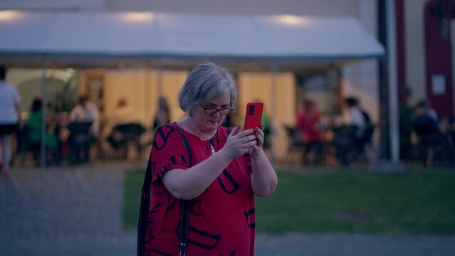 Older woman with grey hair wearing glasses and a red T-shirt taking pictures with her smartphone in the historical city Bardejov in Slovakia during a blue hour at dusk in slow motion. Sony FX3 in 4K