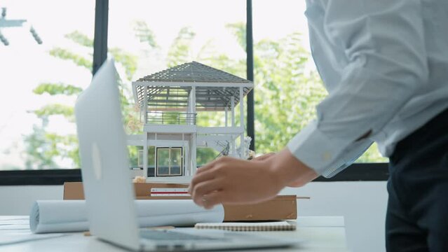 Close up of professional male architect engineer measure house model and using laptop analysis data at meeting table with house model and map . scatter around. Focus on hand. Immaculate.