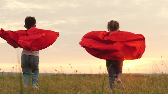 Children running with red cloaks on backs blowing in wind at sunset across field. Children running freely play favourite superheroes at sunset on field. Children dressed as superheroes run to sunset