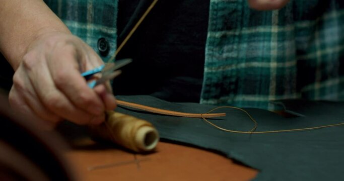 bearded old leather craftsman making handbag. Man with a needle and thread works in leather workshop. Tailor in checked shirt making a purse sewing accessories in atelier. Slow motion