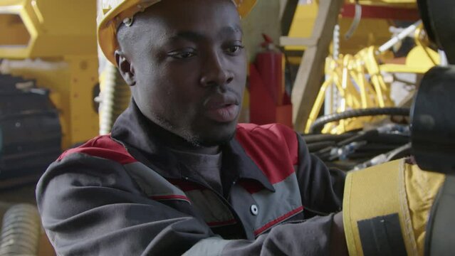 Tilt closeup of African American male workman in yellow hat and uniform fixing elements of internal combustion engine of caterpillar tractor at plant