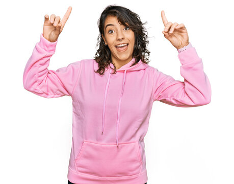 Young hispanic woman wearing casual sweatshirt smiling amazed and surprised and pointing up with fingers and raised arms.