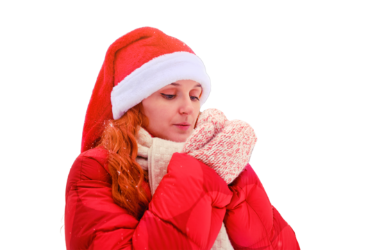 A woman with red hair blows on her frozen hands in knitted mittens, isolated on a white background. Redhead woman on Christmas and New Year Eve walks outdoors in a red hat and jacket