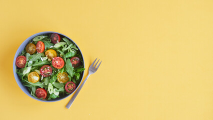 Healthy salad of arugula and red and yellow cherry tomatoes on a bowl next to fork. Top view. Yellow background, copy space.
