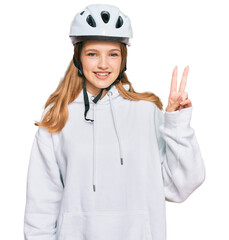 Beautiful young caucasian girl wearing bike helmet smiling with happy face winking at the camera doing victory sign. number two.