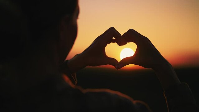 Woman folding hands at sunset in heart shape, symbol of real romantic love, back view. Healthy lifestyle, balance, harmony concept. Female in unity with nature showing heart gesture, enjoying evening.