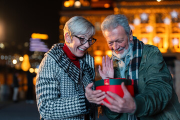 Two senior people looking at gift box while standing outdoors in warm clothing on the street.