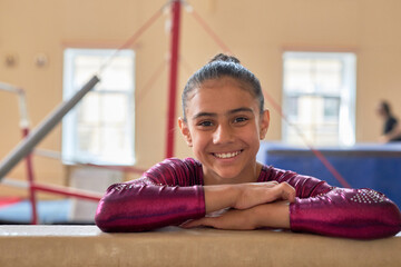 Medium close up portrait of smiling girl in pink gymnastic suit leaning crossed hands on gymnastic...