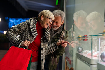 Thrilled senior couple shopping for gifts standing in front of a store at shopping mall. Cheerful elderly man and woman looking for a perfect gift for upcoming holidays.