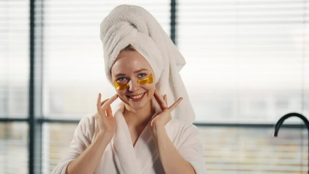 Woman applying hydrogel eye patches. Girl in white bathrobe, towel on head enjoying facial skincare procedure looking at camera smiling in bathroom. Spa, wellness, self care home cosmetology concept.