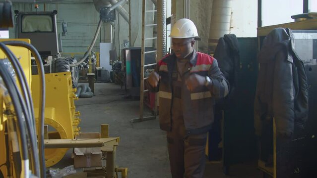 Side shot of African American workman putting on overall jacket and white hard hat and then going to work at machinery plant