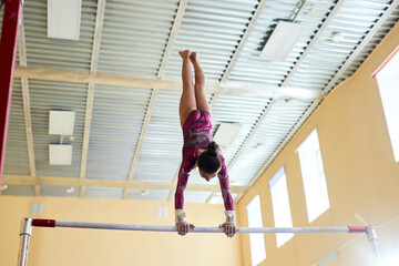 Low angle view at brunette female gymnast dressed in purple leotard pausing in handstand at top of uneven bars, copy space - obrazy, fototapety, plakaty