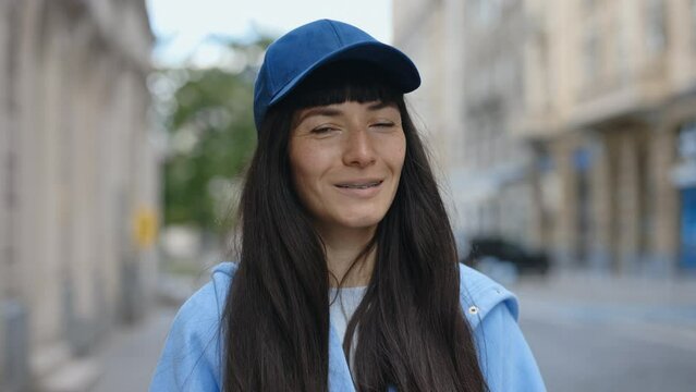 Portrait of Pretty Caucasian Young Lady with Braces in Casual Clothes and Cap Smiling and Looking at Camera while Standing on City Street. Attractive Girl Posing Outdoors. Lifestyle Concept