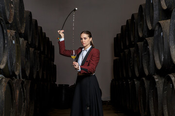 Focused young woman venenciador standing and pouring wine with venencia into glass in cellar