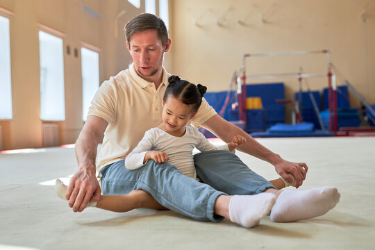 Careful coach sitting on floor with smiling dark-haired little girl gymnast and helping her with leg muscle stretching for performing side splits