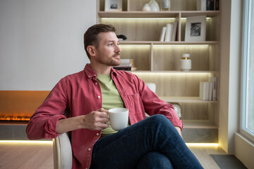 Pensive serious confident pleasant middle-aged unshaved man sitting in cozy comfortable modern apartment, looking at window, drinking tea, thinking, resting, relaxing after long hard working day