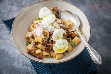 Italienische mini Burrata Käse Kugeln mit Mezzi Rigatoni Pasta, getrockneten Tomaten, Parmesan, Olivenöl, Basilikum, Knoblauch und Chili Flocken in Schale mit Löffel und dunkel grau Stein Hintergrund