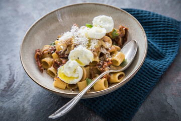 Italienische mini Burrata Käse Kugeln mit Mezzi Rigatoni Pasta, getrockneten Tomaten, Parmesan, Olivenöl, Basilikum, Knoblauch und Chili Flocken in Schale mit Löffel und dunkel grau Stein Hintergrund