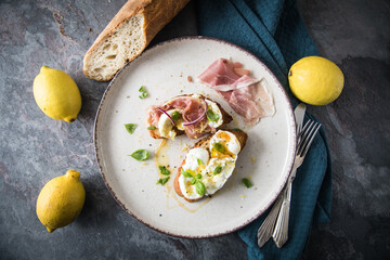 Italienische Burrata Käse Crostini aus Ciabatta Brot,konfierter Knoblauch, Prosciutto Schinken, rote Zwiebel, Basilikum, Honig, Olivenöl, Zitronen mit Teller, Besteck, Serviette blau, dunkel grau Stei