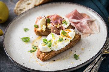 Italienische Burrata Käse Crostini aus Ciabatta Brot,konfierter Knoblauch, Prosciutto Schinken, rote Zwiebel, Basilikum, Honig, Olivenöl, Zitronen mit Teller, Besteck, Serviette blau, dunkel grau Stei