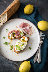 Italienische Burrata Käse Crostini aus Ciabatta Brot,konfierter Knoblauch, Prosciutto Schinken, rote Zwiebel, Basilikum, Honig, Olivenöl, Zitronen mit Teller, Besteck, Serviette blau, dunkel grau Stei