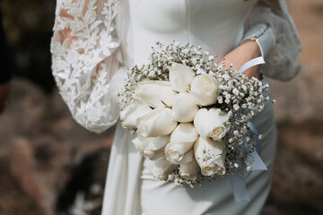 The bride in a long tight white wedding dress holds a beautiful bouquet of white roses in her hands during the day. Image for your design or creative compositions. - obrazy, fototapety, plakaty