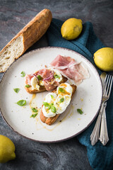 Italienische Burrata Käse Crostini aus Ciabatta Brot,konfierter Knoblauch, Prosciutto Schinken, rote Zwiebel, Basilikum, Honig, Olivenöl, Zitronen mit Teller, Besteck, Serviette blau, dunkel grau Stei