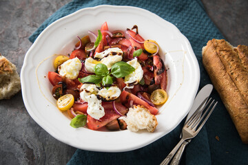 Italienischer Burrata Käse Salat mit bunten roten und gelben Tomaten, Basilikum, Ciabatta Brot Olivenöl und Balsamico Dressing mit tiefer Teller, Besteck Serviette blau, dunkel grau Stein Hintergrund