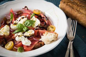 Italienischer Burrata Käse Salat mit bunten roten und gelben Tomaten, Basilikum, Ciabatta Brot Olivenöl und Balsamico Dressing mit tiefer Teller, Besteck Serviette blau, dunkel grau Stein Hintergrund