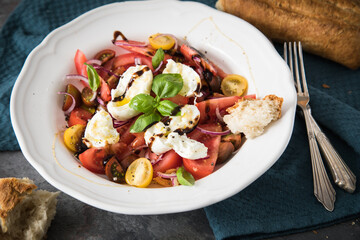 Italienischer Burrata Käse Salat mit bunten roten und gelben Tomaten, Basilikum, Ciabatta Brot Olivenöl und Balsamico Dressing mit tiefer Teller, Besteck Serviette blau, dunkel grau Stein Hintergrund