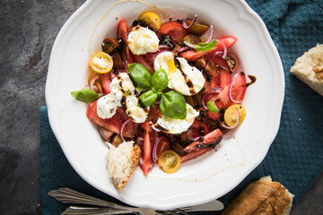 Italienischer Burrata Käse Salat mit bunten roten und gelben Tomaten, Basilikum, Ciabatta Brot Olivenöl und Balsamico Dressing mit tiefer Teller, Besteck Serviette blau, dunkel grau Stein Hintergrund